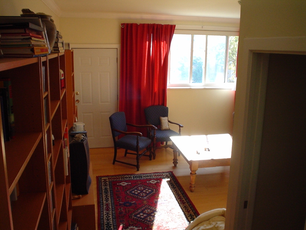 Ein Zimmer mit einem Teppichboden, einem Bücherregal links, einem Tisch mit zwei Stühlen rechts, Glasfenstern mit roten Vorhängen im Hintergrund, einer Tür neben den Vorhängen, einer Maschine links und einer weiteren Tür unten rechts.