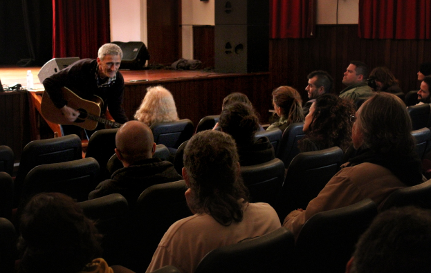 Ein älterer Mann in einem schwarzen Pullover spielt Gitarre auf einer Bühne mit Lautsprechern und einem Vorhang, während viele Menschen auf Stühlen sitzen und ihm zuschauen.
