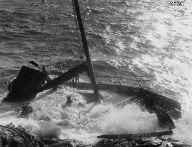 Ein Schwarz-Weiß-Foto eines beschädigten Bootes auf dem Meer, dessen Segel flattern und dessen Rumpf teilweise unter Wasser ist.