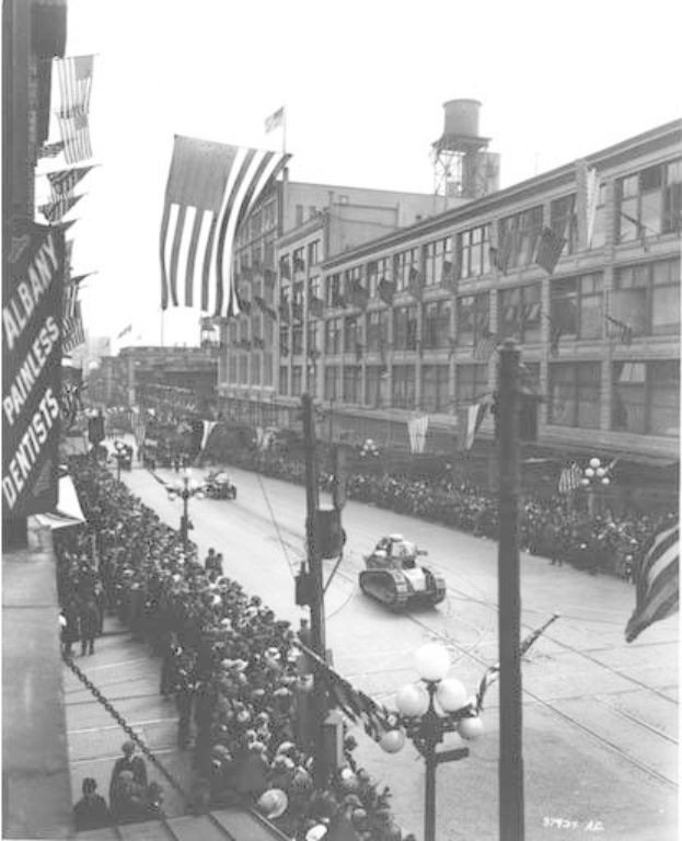 Schwarze und weiße Fotografie einer Menge, die eine Straße entlang geht, während ein Umzug in Albany, New York City, USA am 4. Juli 1939 stattfindet, mit Fahrzeugen, Laternenmasten, Gebäuden und einem klaren Himmel im Hintergrund.