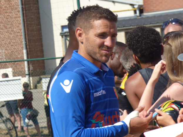 Ein Mann in einem blauen Hemd signiert Autogramme für eine Gruppe von Menschen, einige tragen Kappen und Schutzbrillen, mit einem Gebäude, Maschendrahtzaun und einem Fahrrad im Hintergrund.