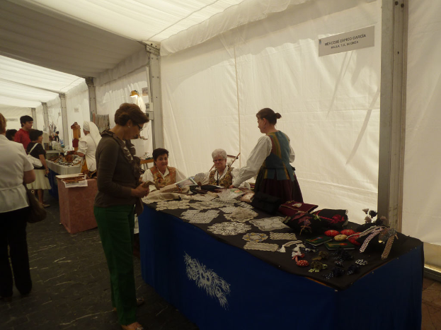 Ein vielbesuchtes Ereignis mit verschiedenen Ständen, Menschen, die stehen und sitzen, Tätowierungen und Gegenständen auf einem Tisch im Vordergrund und weißgekleideten Figuren im Hintergrund.