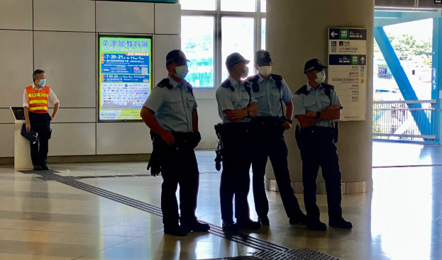 Eine Gruppe von Polizisten in Masken steht vor einem Gebäude mit Glastüren, Schildern mit Text, einer Säule und einem Geländer, was darauf hinweist, dass sie Personen in Hong Kong festgenommen haben.