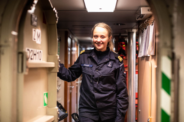 Eine Frau in schwedischer Polizeiuniform lächelt in einem Flur mit nummerierten Türen auf der linken Seite und Papieren auf der rechten Seite, mit einer Deckenlampe im Hintergrund.
