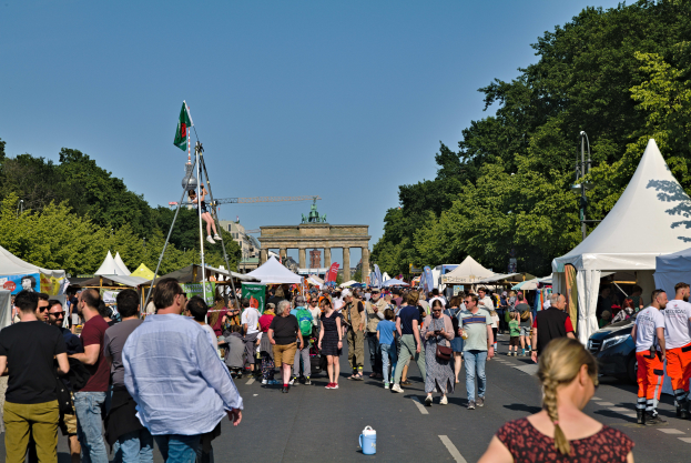 Eine belebte Straßen Szene mit Menschen, die an Zelten, Fahrzeugen und Bäumen vorbeigehen, unter einem klaren blauen Himmel mit einem Bogen in der Ferne und Fahnenmasten auf der linken Seite, wahrscheinlich das Oktoberfest in München, Deutschland.