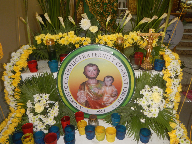 Ein Tisch geschmückt mit farbenfrohen Blumen, Kerzen und einem Schild mit einem Porträt und Text, vor einem Hintergrund aus Pflanzen, Stühlen und anderen Gegenständen, die eine festliche Atmosphäre für die Bruderschaft von Sankt Joseph schaffen.