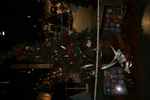 Ein Weihnachtsbaum mit Geschenken, Rahmen und Spielzeugen auf einer Plattform, mit Menschen, weiteren Weihnachtsb├Ąumen, einer Kette, einem Pfahl und Lichtern im Hintergrund.