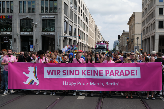 Eine Gruppe von Menschen, die eine Straße in Berlin, Deutschland, entlanggehen und ein pinkes Banner mit der Aufschrift "Happy Pride March" halten, mit Gebäuden, Laternenmasten und Verkehrszeichen an der Straße, unter einem bewölkten Himmel.