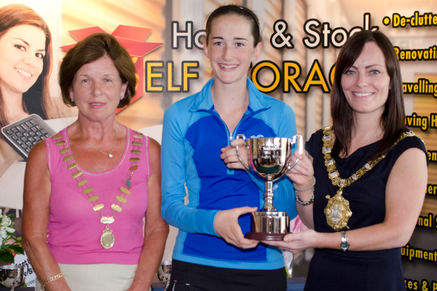 Drei Frauen stehen zusammen, wobei eine eine pinkfarbene Bluse trägt, eine eine blaue T-Shirt mit einem Becher hält und eine in einem schwarzen Kleid den Becher präsentiert, mit einem Banner im Hintergrund mit Bildern und Text.