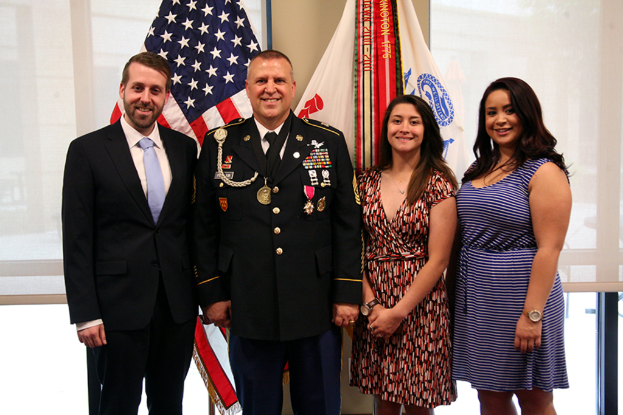 Zwei Frauen in Kleidern und ein Mann in einem Anzug, die drinnen stehen und lächeln, mit einer Person in Uniform, die ebenfalls lächelt, Jalousien und Fahnen im Hintergrund.