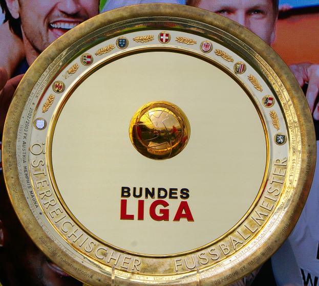 Ein Mann hält ein goldenes Schild mit der Aufschrift 'Bundesliga' hoch, im Hintergrund ist eine Fahne mit drei Personen zu sehen.