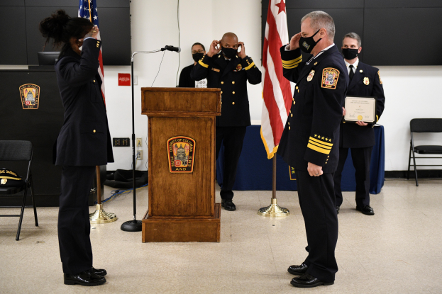 Gruppe uniformierter Personen in Masken vor einem Podium mit Mikrofon, umgeben von Fahnen und Stühlen, während einer Vereidigung eines neuen Feuerwehrchefs, wobei eine Person ein Zertifikat hält.