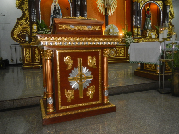 Ein Altar in einer Kirche mit einem Kreuz, Statuen, Blumenbouquets, Kerzen, einem tischdecke-bedeckten Tisch und anderen Gegenständen, an den Wänden mit Lichtern geschmückt.
