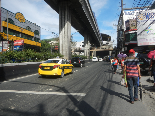 Gelbes Taxi fährt eine Stadtstraße mit hohen Gebäuden, Fußgängern auf dem Gehweg mit Regenschirmen, einer Brücke im Hintergrund, bewölktem Himmel und Strommasten mit Oberleitungen.