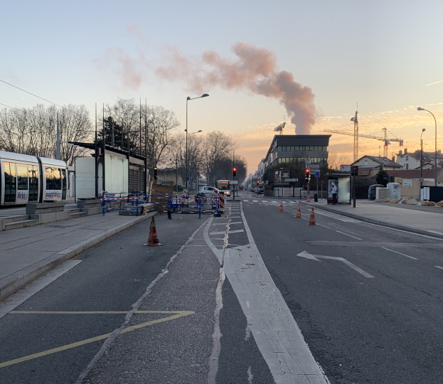 Stra√enszene mit Verkehrskegeln, einem Bus, Laternenpfählen, Bäumen, Gebäuden, Kränen und Rauch, der aus einem entfernten Gebäude aufsteigt, vor einer Stadtkulisse.