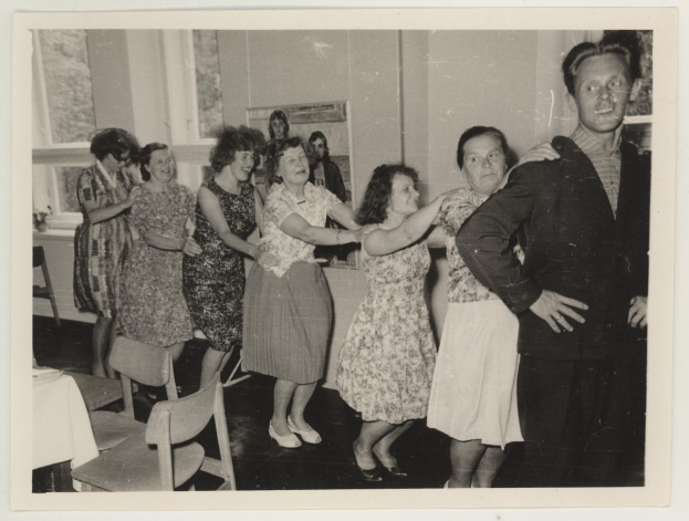 Eine Gruppe von Menschen, die in einem Raum im Stil der 1950er Jahre tanzen, mit Stühlen, einem Tisch mit einer wei├čen Decke, einem Foto auf der Wand und Fenstern im Hintergrund.