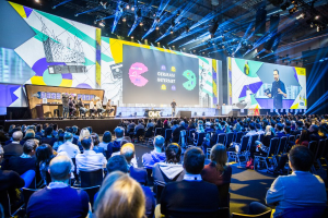 Eine große Menschenmenge, die auf Stühlen vor einer Bühne sitzt, mit einem Tisch voller Gegenstände links und Bildschirmen und Lichtern im Hintergrund.