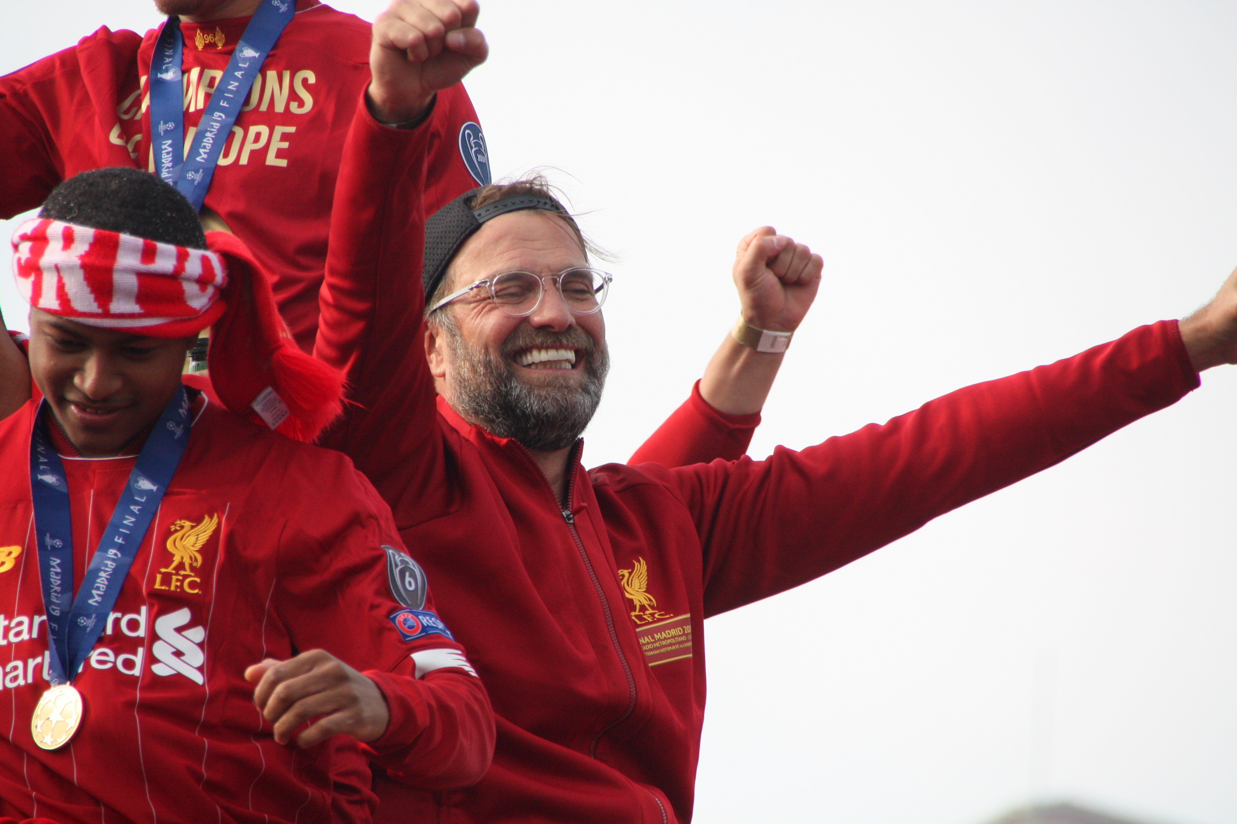 Liverpool-Trainer Jürgen Klopp feiert mit seinen Spielern den UEFA-Champions-League-Sieg, während zwei medaillentragende Personen in der Nähe sind und der Himmel im Hintergrund zu sehen ist.