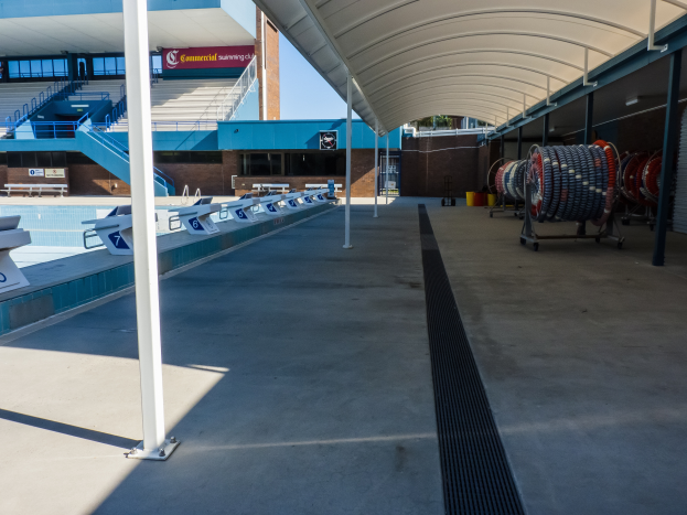 Eine große Hallen-Schwimmhalle an der University of Texas at Austin mit Stühlen drum herum, Gegenständen auf dem Boden rechts, Pfählen links und einem Gebäude mit Treppen, Geländern und Texttafeln im Hintergrund.