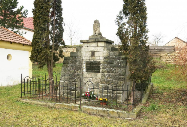 Ein steinernes Denkmal auf einer grünen Wiese, umgeben von einem Metallzaun und geschmückt mit Blumensträußen, mit Bäumen, Häusern und einer Wand im Hintergrund unter einem klaren blauen Himmel dient als Holocaust-Gedenkstätte in Sibiu, Rumänien.
