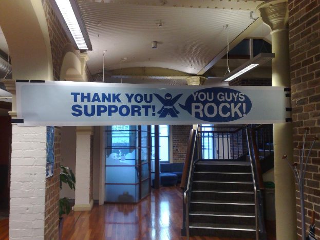 Ein Dankeschön-Banner hängt an einer Wand und einer Säule vor einer Treppe, mit Fenstern im Hintergrund.