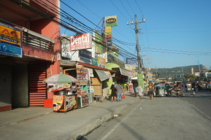 Eine belebte Stadtstraße mit Fußgängern, Gebäuden, Geschäften, Schildern, Strommasten, Straßenlaternen, Bäumen und Hügeln unter einem klaren blauen Himmel.