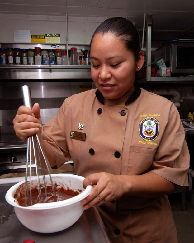 Eine Frau in Kochuniform steht in einer Küche und hält einen Topf mit einem Schneebesen in der Hand, mit verschiedenen Gegenständen auf dem Tisch und Flaschen auf Gestellen im Hintergrund.