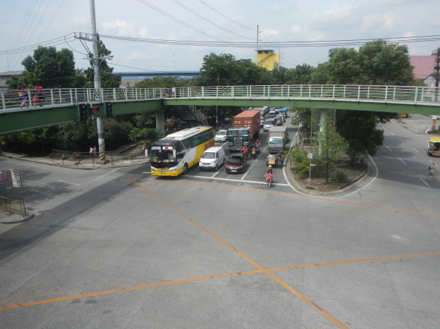Eine belebte Straße mit Autos und Bussen unter einer Brücke, Menschen auf der Brücke, Strommasten mit Kabeln, Bäume, Gebäude und ein bewölkter Himmel im Hintergrund.