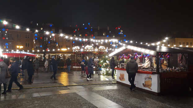 Ein belebter Weihnachtsmarkt bei Nacht in einer Stadt mit Menschen, geschmückten Ständen, Gebäuden, Bäumen und festlichen Lichtern am Himmel.