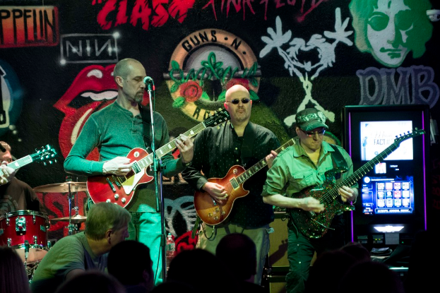 Drei Männer auf einer Bühne mit Gitarren, umgeben von einer Menge und einer bemalten Wand im Hintergrund.