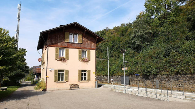 Ferienhaus zum Verkauf in Baden-Württemberg, Deutschland, mit einem mehrfenstrigen Gebäude mit einer vorderen Einfriedung, umgeben von Straßeninfrastruktur, Grünflächen und einem bewölkten Himmel.