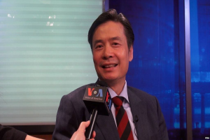 Ein Mann im Anzug hält ein Mikrofon, mit der Hand eines Reporters sichtbar, der vor einem Fenster und einer Wand spricht.