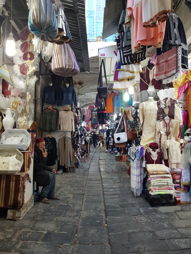 Ein belebter Straßenmarkt in Jerusalem Altstadt mit verschiedenen Artikeln wie Kleidung, Taschen und Lichter auf dem Marktplatz und Menschen, die die Stände durchstöbern, mit einem Gebäude im Hintergrund.