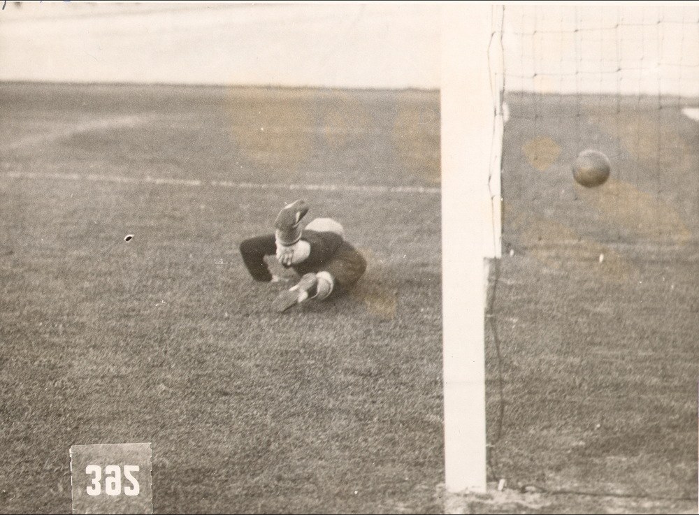 Schwarzes und weißes Foto eines Fußballspielers in Fußballschuhen, der einen Ball in der Nähe eines Torpfostens mit Netz auf der rechten Seite fängt, mit Text unten.