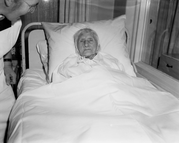 Ein älteres Frau liegt in einem Krankenhausbett mit einer weißen Decke und Kissen, während eine Krankenschwester neben ihr steht, mit einem Fenster und Vorhängen im Hintergrund.