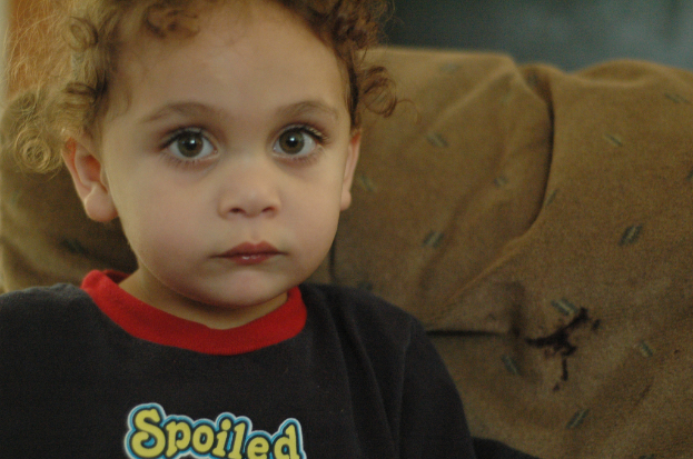 Ein Kind mit lockigen Haaren, das ein schwarzes Hemd mit der gelben Aufschrift "Spoiled" trägt, vor einem braunen Stoffhintergrund.