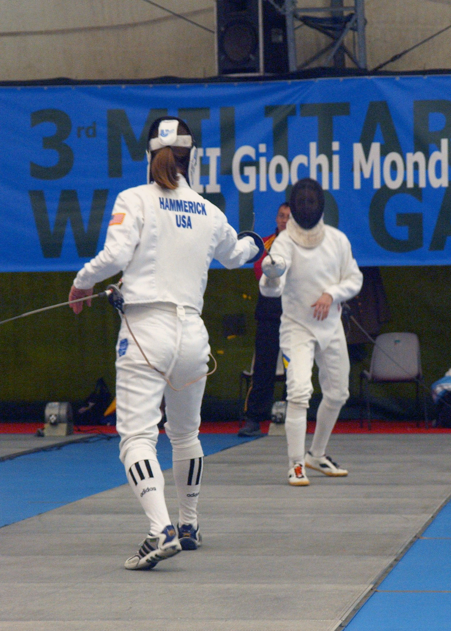 Zwei Fechter in weißen Kostümen und mit Schwertern gegenüberstehen bei einem olympischen Event, mit einem blauen Banner und Zuschauern im Hintergrund.
