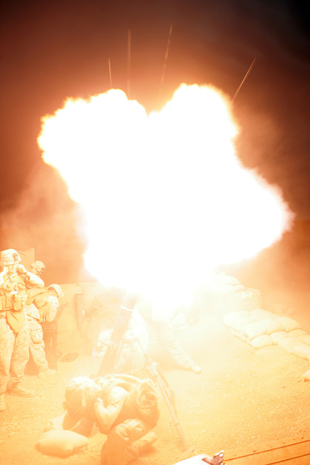 Mehrere Personen in Uniformen und Helmen befinden sich auf einem Gelände mit ein paar umherliegenden Taschen, wobei in der Mitte der Szene ein Feuer brennt.