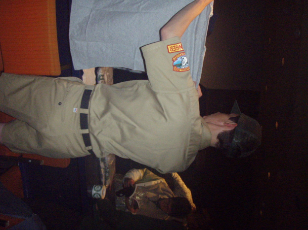 Ein Polizeibeamter hält ein T-Shirt in den Vordergrund, mit einem Jungen im Hintergrund, der ein Buch und einen Becher hält und in der Nähe eines Schreibtischs und Lampen steht.