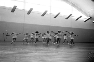 Eine Gruppe junger Mädchen, die sich an den Händen halten und in einem schwarz-weißen Tanzstudio tanzen, mit einem Ball auf der rechten Seite und Lampen an der Decke.