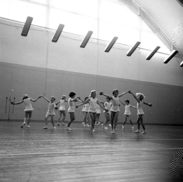Eine Gruppe junger Mädchen, die sich an den Händen halten und in einem schwarz-weißen Tanzstudio tanzen, mit einem Ball auf der rechten Seite und Lampen an der Decke.