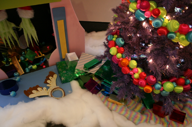 Innen: Ein geschmückter Weihnachtsbaum rechts, Geschenkboxen und Dekorationen in der Mitte, eine Hütte mit Dekorationen oben links und Baumwolle und Wolle unten.