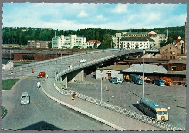 Altes Schwarz-Weiß-Foto einer Stadtstraße mit Fahrzeugen, Fußgängern auf einer Brücke, Gebäuden, Bäumen, Laternen und einem bewölkten Himmel.