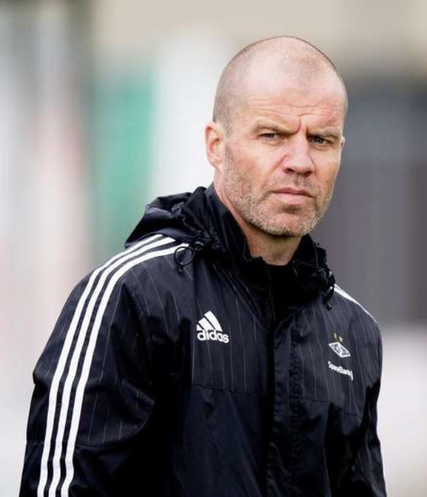 Baldiger Trainer in marineblauer Jacke mit weißen Streifen und weißem Kragen auf einem Fußballfeld stehend, Hände verschränkt, mit ernster Miene und unscharfem Hintergrund.