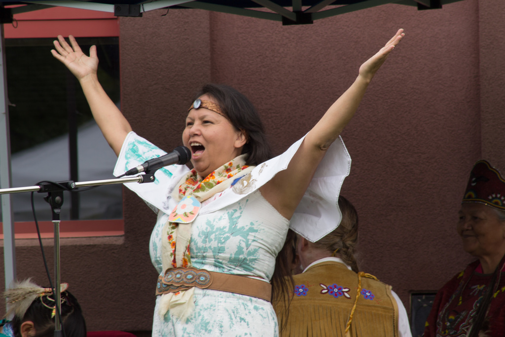 Eine Frau in einem weißen Kleid steht auf einer Bühne, schreiend in ein Mikrofon, mit einer Wand und einem Eisenrahmen dahinter.