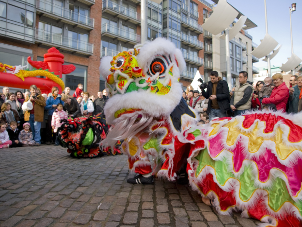 Lebendige Feier des chinesischen Neujahrs in Amsterdam mit einem Löwen Tanz im Vordergrund und einer Menge Menschen drumherum, einige halten Kameras, vor einem Hintergrund aus Gebäuden, Laternenmasten und einem klaren blauen Himmel.