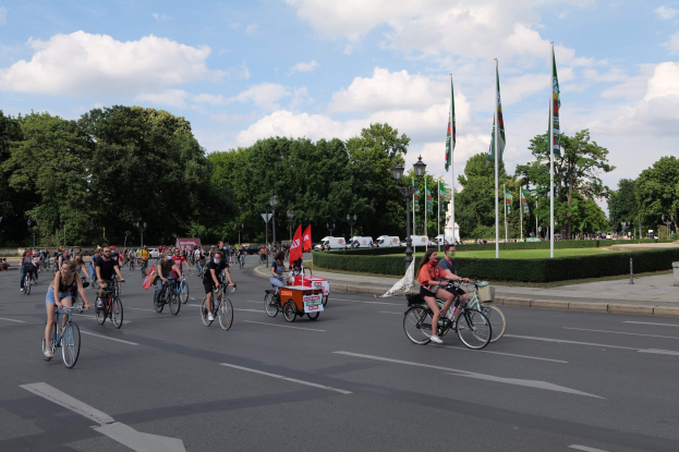 Eine Gruppe von Menschen, die auf Fahrrädern eine von Bäumen gesäumte Straße mit Laternenmasten und Flaggenmasten entlangfahren, während der Pariser Radtour 2016, mit im Hintergrund sichtbaren Fahrzeugen und einem bewölkten Himmel.