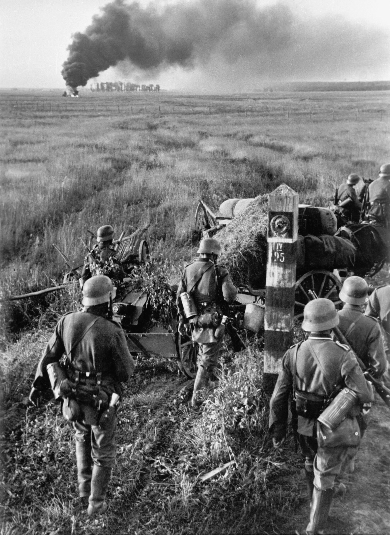 Schwarzes und weißes Bild deutscher Soldaten in Helmen mit Gewehren neben einer Kanone auf einem grasbewachsenen Feld mit Rauch im Hintergrund und Himmel darüber.