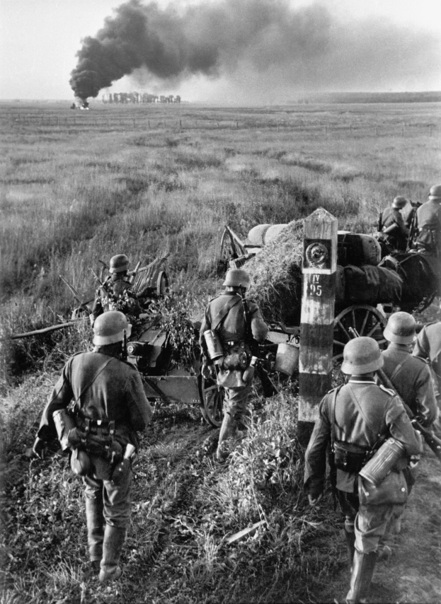 Schwarzes und weißes Bild deutscher Soldaten in Helmen mit Gewehren neben einer Kanone auf einem grasbewachsenen Feld mit Rauch im Hintergrund und Himmel darüber.