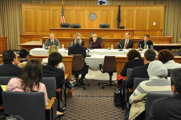 Ein Versammlungsraum mit einer Gruppe von Menschen, die auf Stühlen sitzen, zwei Flaggen anderer Länder und ein orangefarbener Schreibtisch mit Wasserflaschen und einem Mikrofon.